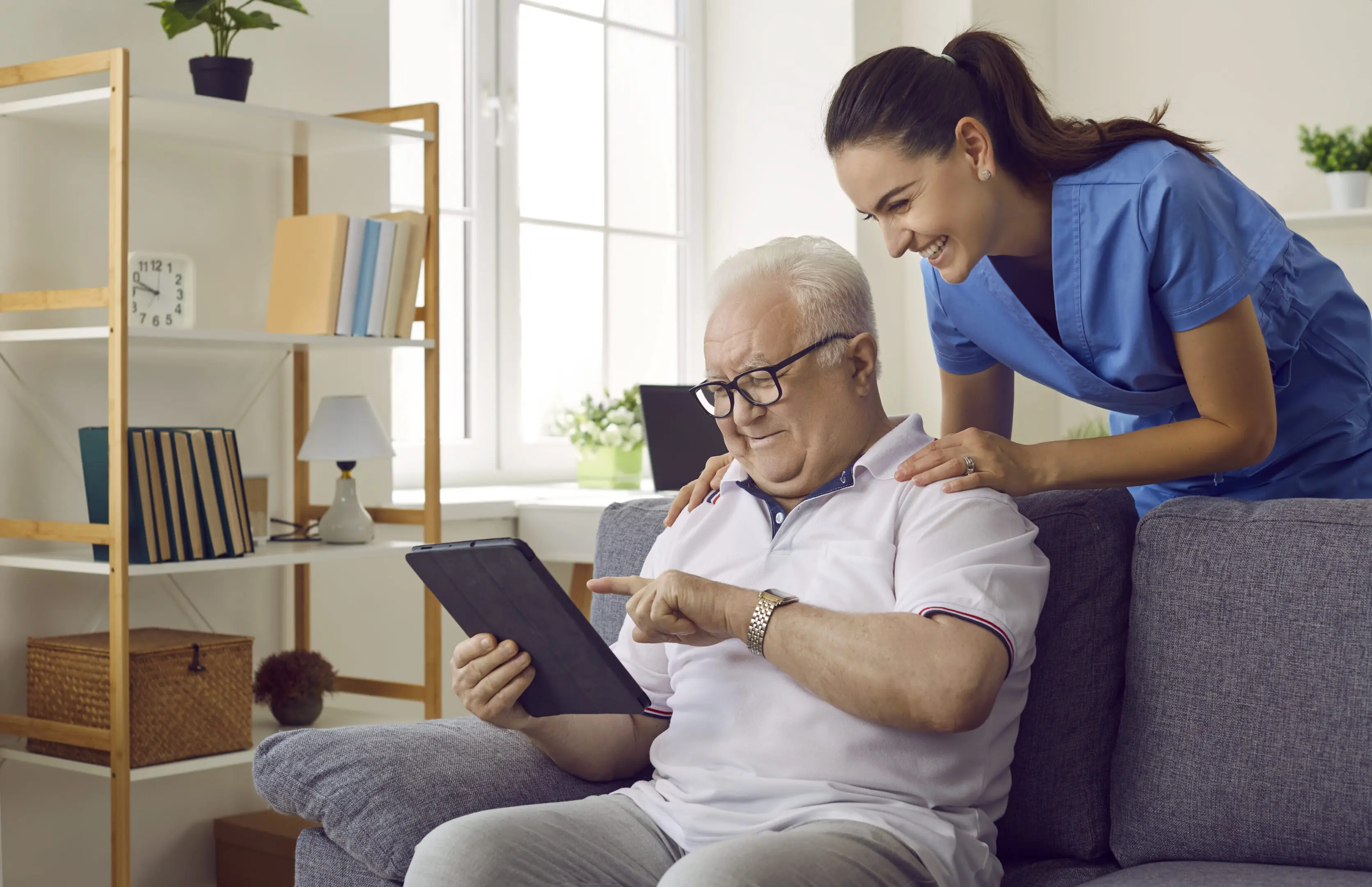 Nurse shows old man how to use apps on digital tablet to monitor his health and how to read e-books. Smiling pensioner looking at screen of modern device. Elderly people healthcare tech concept.